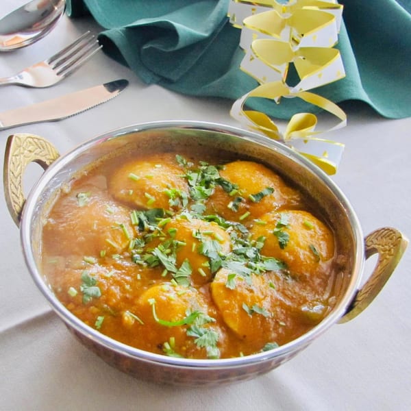 A metal bowl of Indian egg curry with cilantro, silverware, and a green napkin on a white tablecloth.