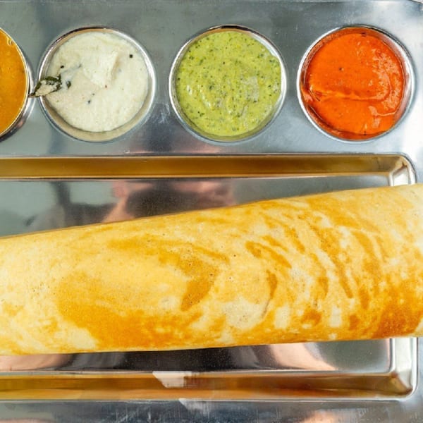 A golden-brown dosa on a metal tray, with four small bowls of colorful chutneys and sambar.