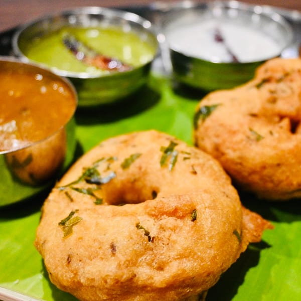 Two golden-brown Medu Vada on a banana leaf, served with three chutneys in steel bowls.